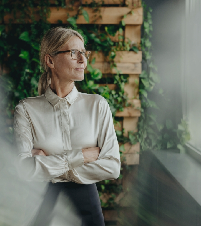 Mature woman wearing work attire and glasses stands in front of a wall of plants and looks confidently out an office window